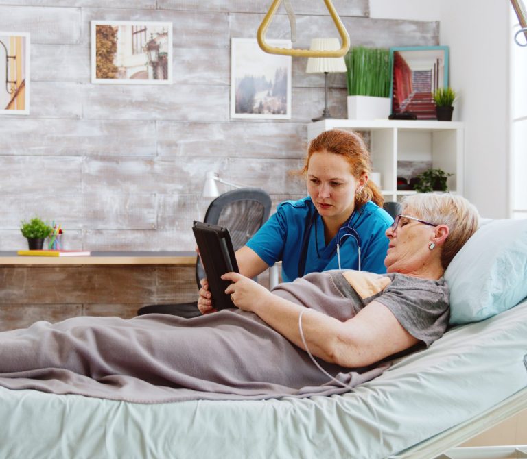 female-caregiver-helps-old-disabled-woman-lying-hospital-bed-use-digital-tablet-pc-bright-room-with-big-windows